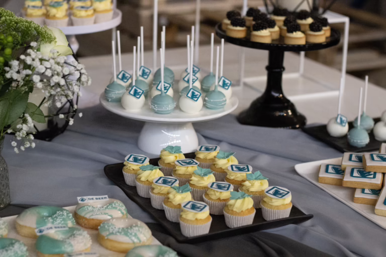 Zakelijke sweet table in zwart-turquoise- branding kleuren. Op veel gebakjes logo decoratie. Donut wall, macaron toren, branded cupcakes - prachtig gepresenteerd.